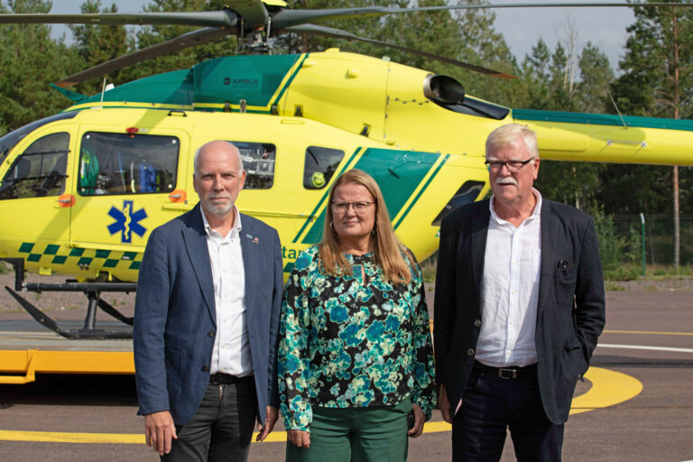 Anders Öberg, ordförande, Anna-Lena Danielsson, vice ordförande och Ulric Andersson, ledamot framför en gul helikopter, efter en presskonferens vid SLAs 10-årsjubileum.
